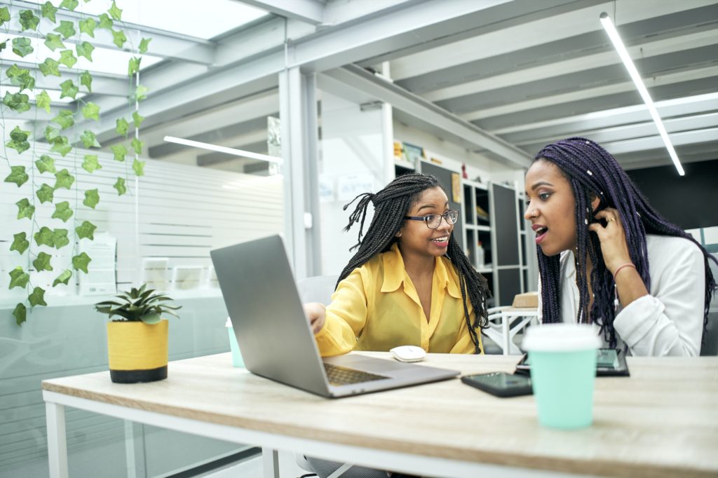 Two young women in office looking surprised at something they are seeing on laptop screen.
